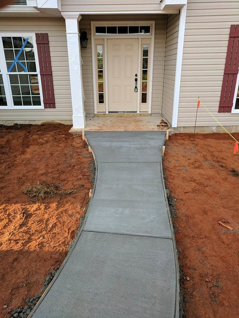 Curved concrete walkway leading to a white door with glass panels and black handle, blue tape visible on adjacent window, construction materials present near entry porch