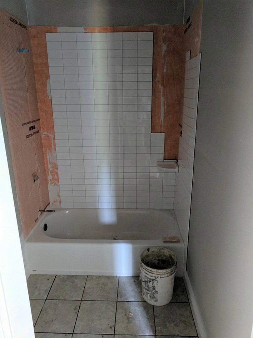 Bathroom with white tile walls, grey tile floor, freestanding bathtub, metal bucket beside tub, and sink visible in background