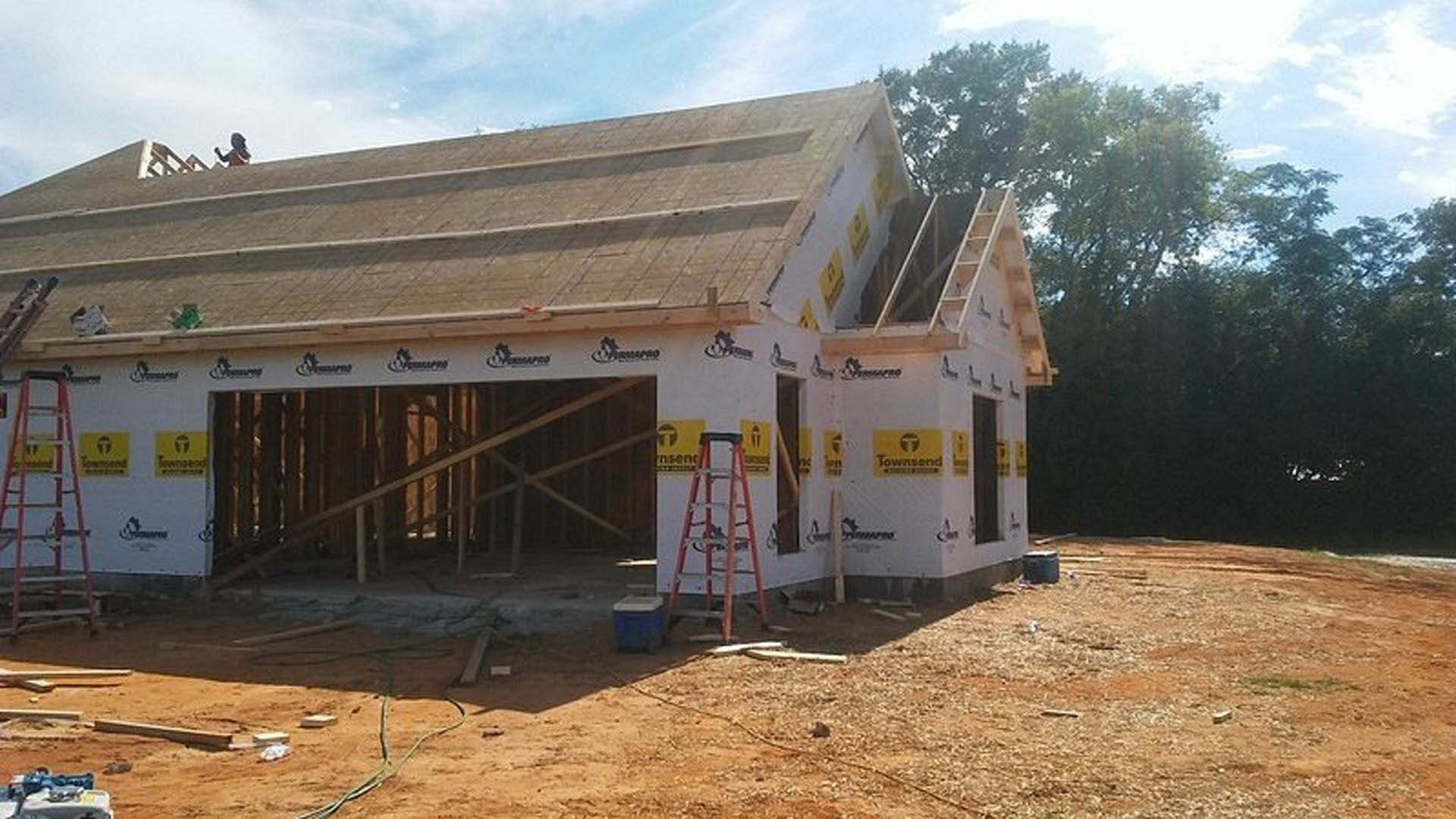 Framed house under construction with exposed wooden walls, red ladder leaning against structure, blue and white cooler on ground, partially built wooden railing, cloudy sky and