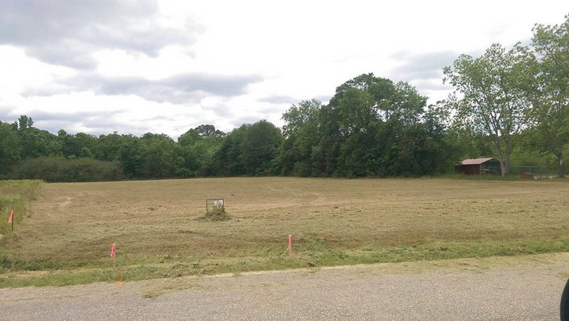 Grassy field bordered by trees under a cloudy sky, red pole and small wooden box in foreground, rural land lot with sign on building in background