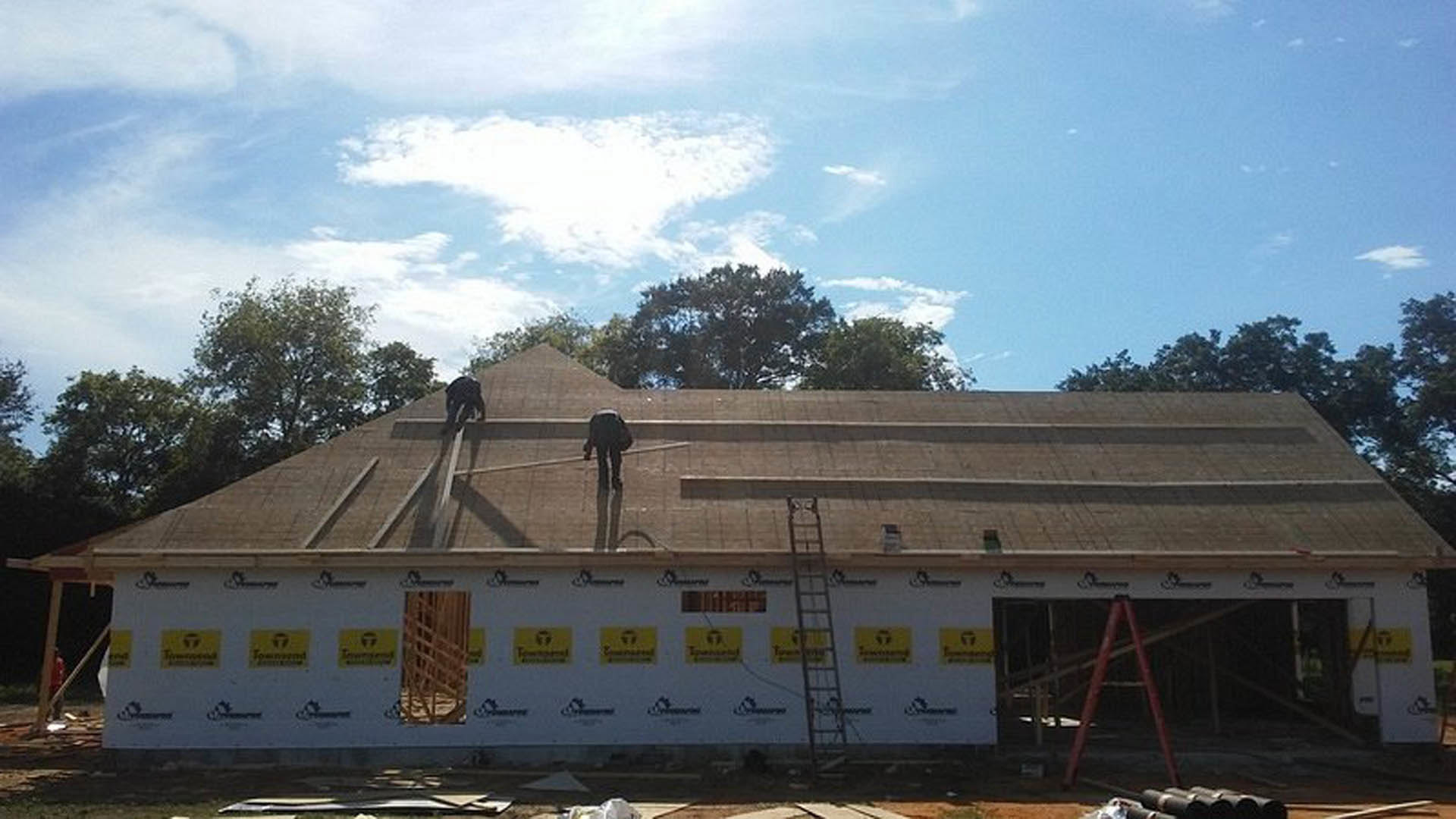 Framed residential structure with exposed wooden beams, workers installing roofing materials, surrounded by trees under a cloudy sky