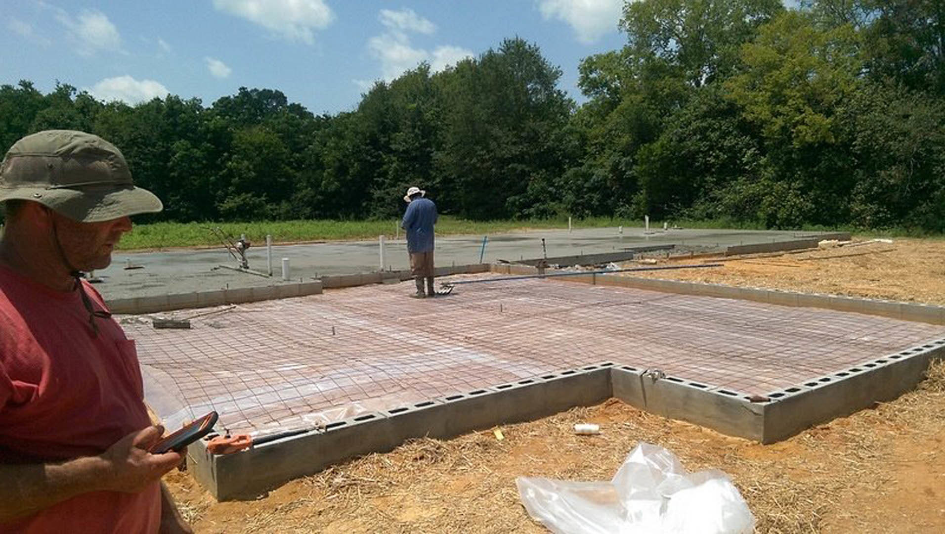 Man in a hat holding a tablet, standing on a newly poured concrete slab outdoors with trees and cloudy sky in background