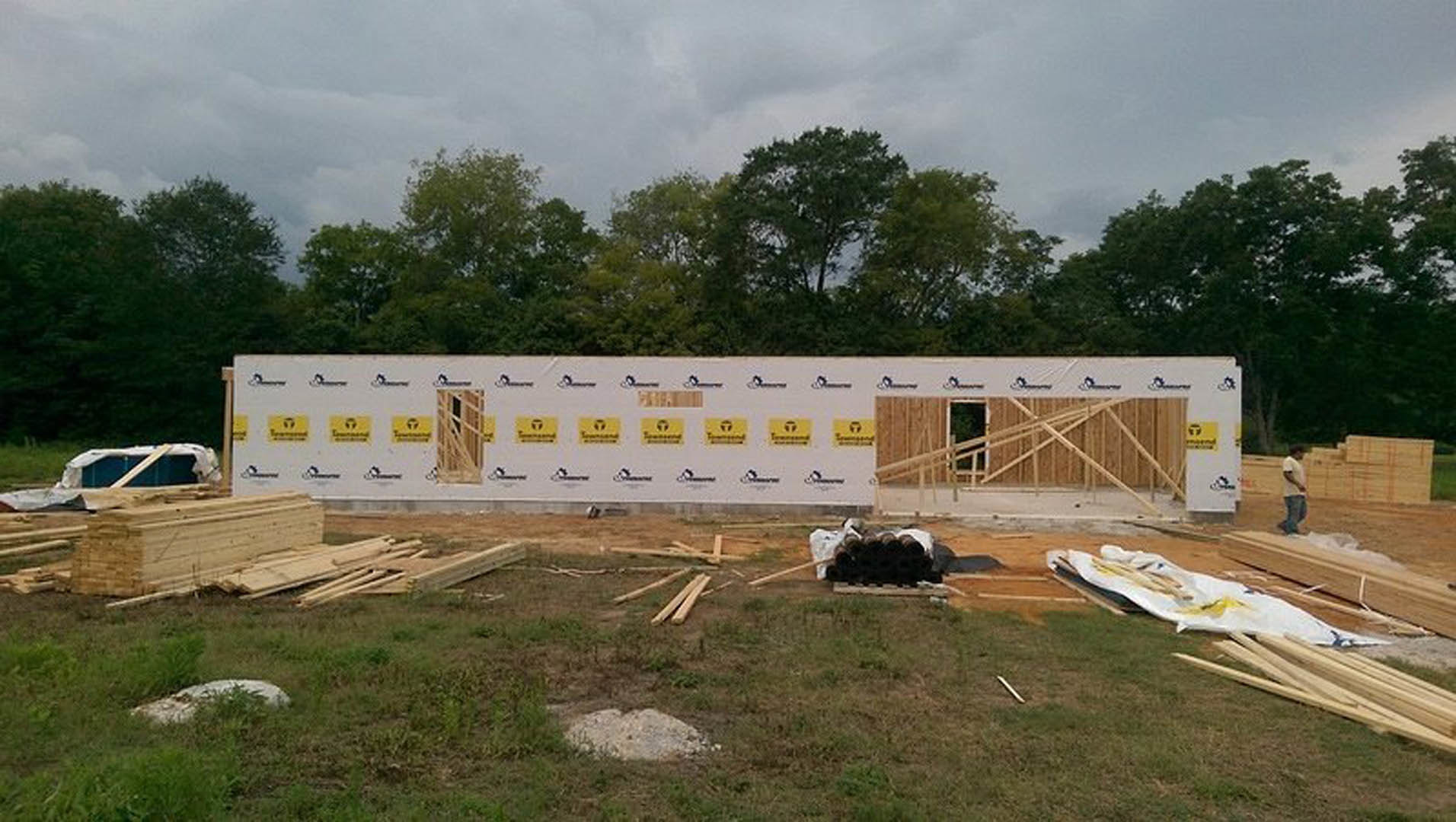 Framed custom home under construction with exposed wooden beams, stacks of lumber in foreground, white sign with yellow stickers, grassy lot bordered by trees under cloudy sky
