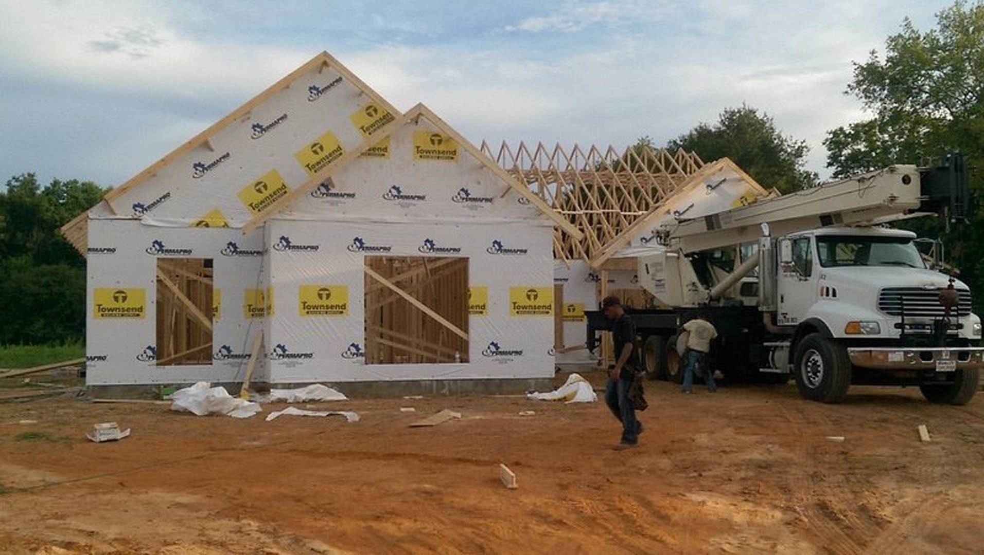 Framed house under construction with exposed wood beams, construction vehicles parked nearby, dirt ground, and cloudy sky overhead
