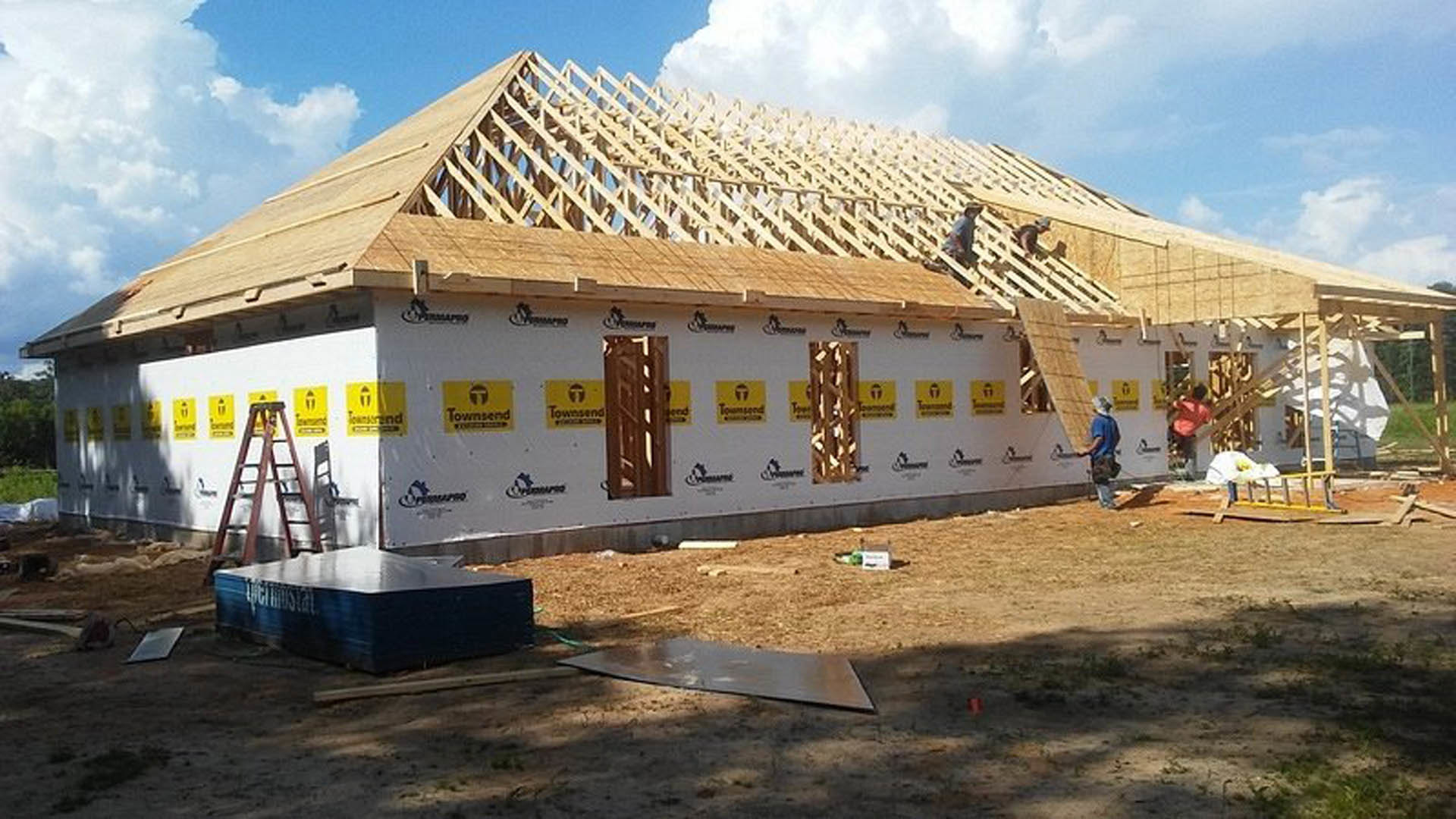 Wood-framed house under construction with exposed beams, wet rectangular object on ground, yellow sign with black text, cloudy sky in background, blurry person near structure