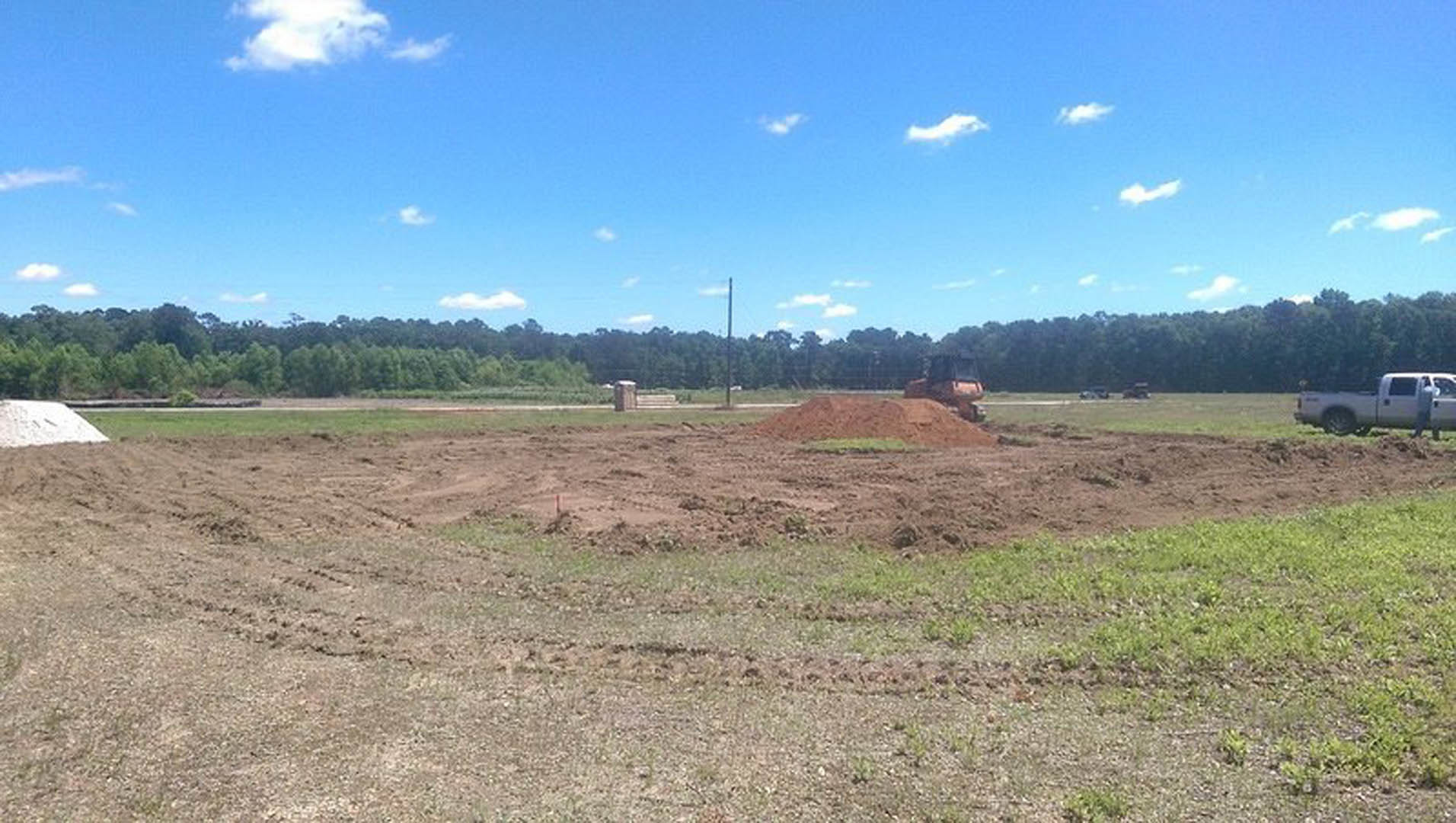 Dirt field with scattered piles of soil, white truck parked near a black door, tractor in the background, blue sky with white clouds overhead, rural landscape with sparse