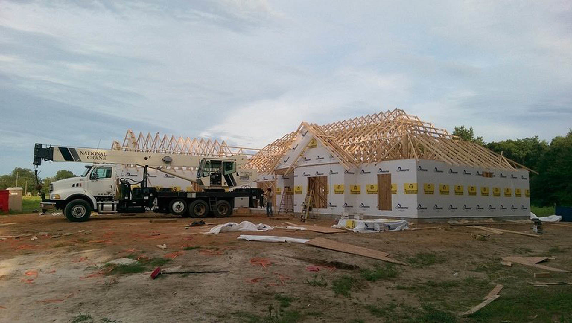 Framed house under construction with exposed wooden beams, partially completed roof, construction crane truck, scattered lumber, and cloudy sky in background