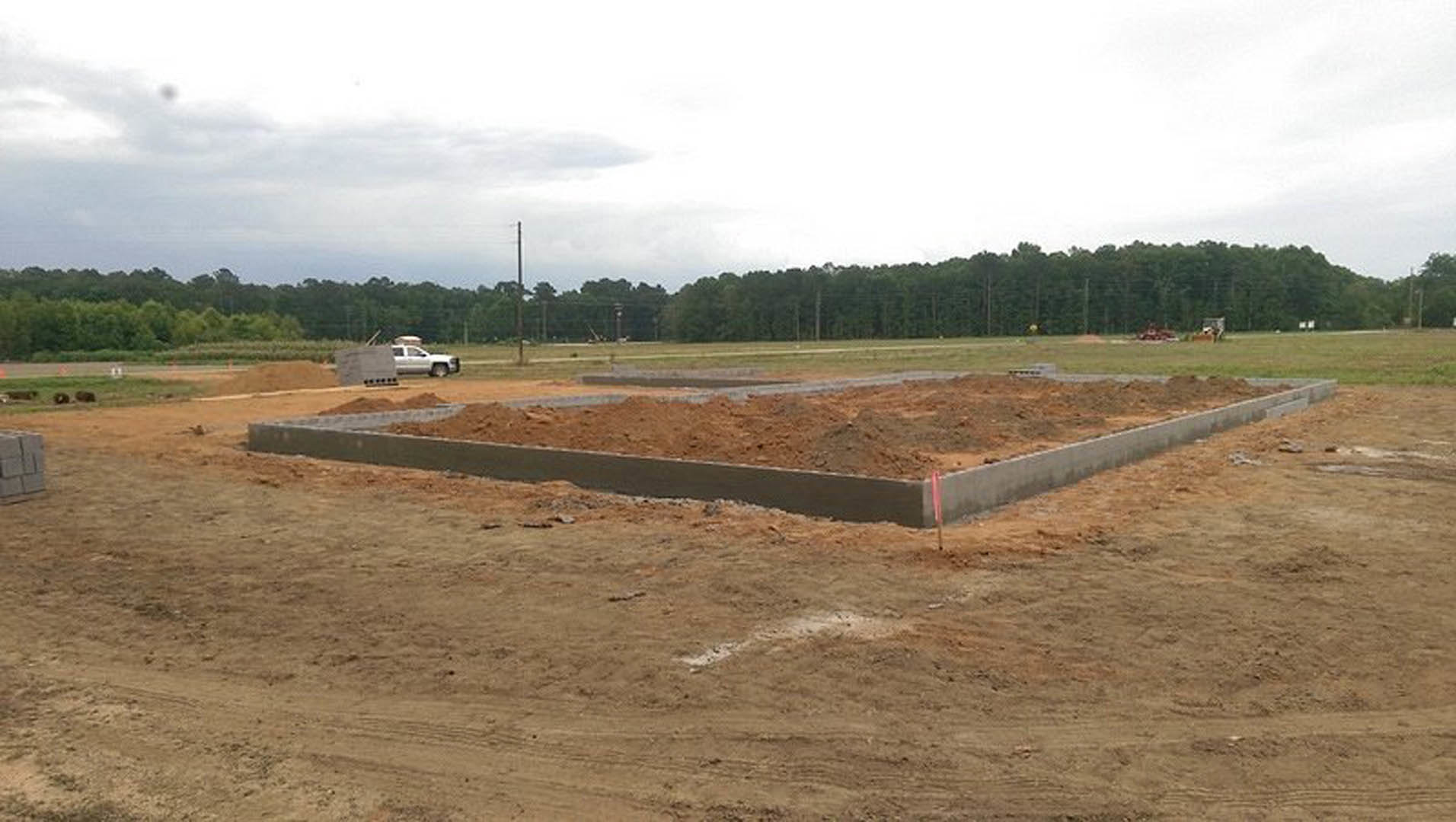 Concrete foundation and dirt lot with white truck parked beside construction site, cloudy sky overhead, grassy field and trees in background.