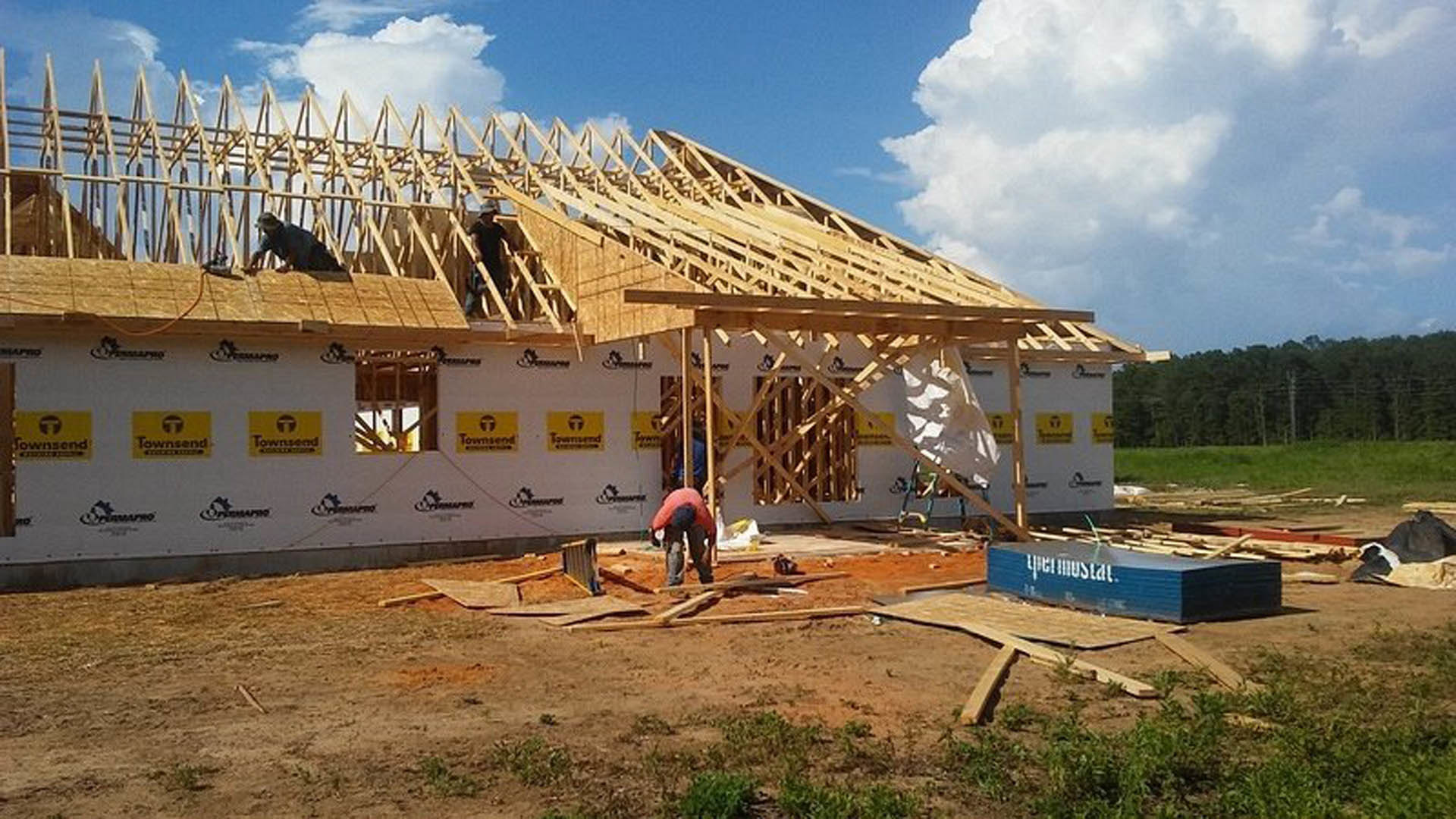 Wood-framed house under construction with workers installing roof panels, surrounded by dirt ground and scattered trees under a cloudy sky
