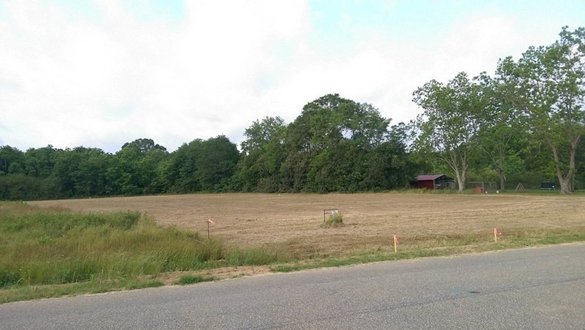 Grassy field bordered by a wire fence, scattered trees in the background, small shed near the edge, rural road running alongside, cloudy sky overhead