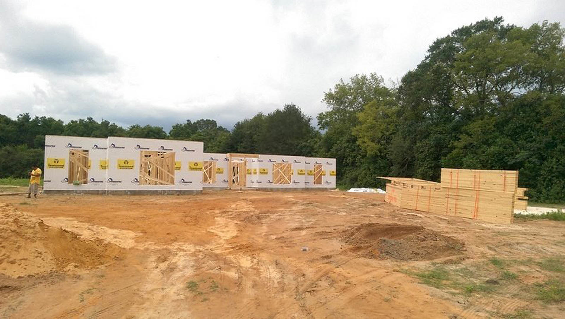 Wood-framed custom home under construction on a dirt lot, surrounded by trees and scattered lumber, cloudy sky overhead