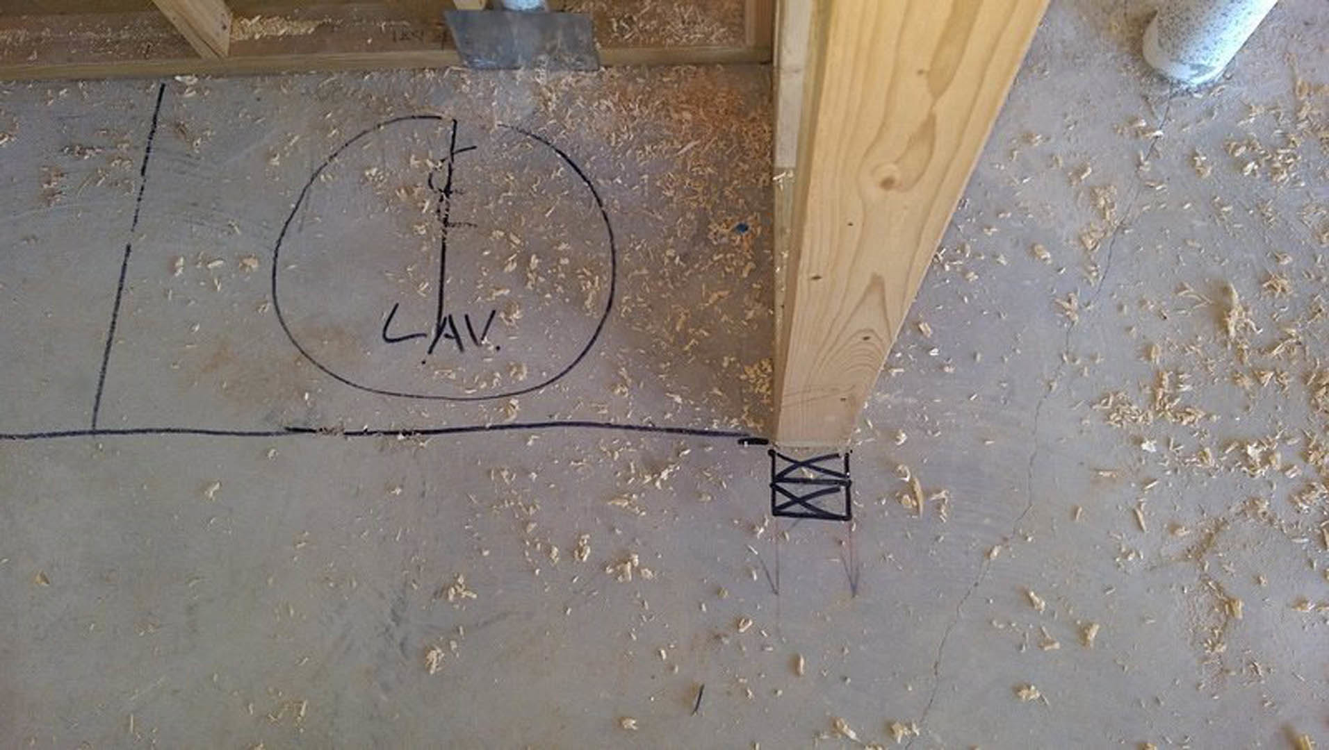 Wood beam resting on plywood floor with black marker handwriting, circular object with symbol, hammer, and black and white box nearby.