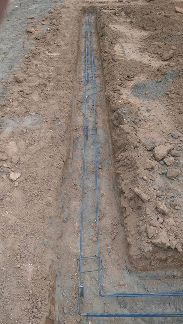 Narrow dirt trench outdoors with exposed electrical wires running along soil and concrete, surrounded by rocks and metal bars