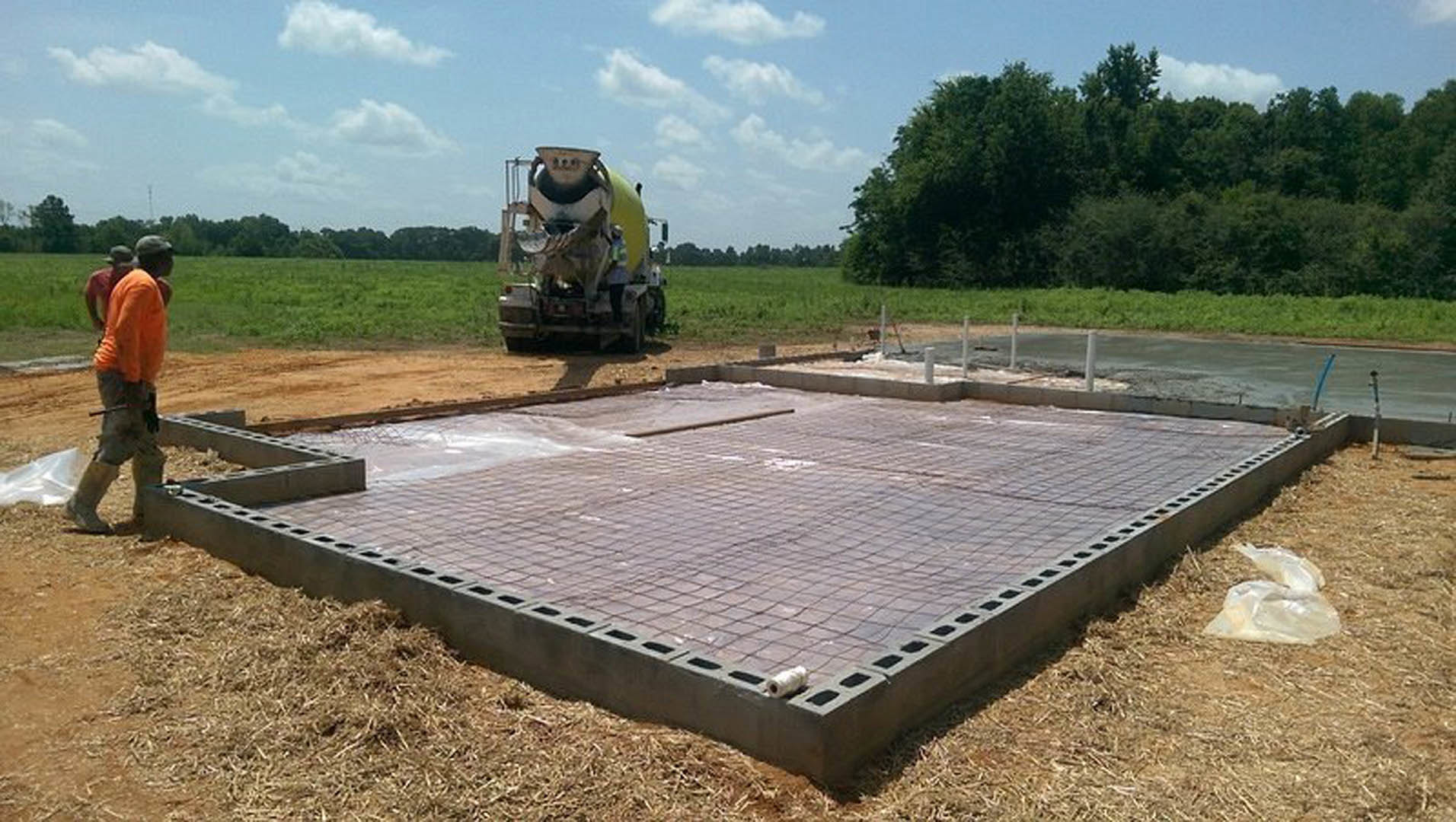 Concrete foundation slab with a white roll of material on the ground, cement mixer and person in boots nearby, blurry cow on a truck in the background, trees and sky visible.