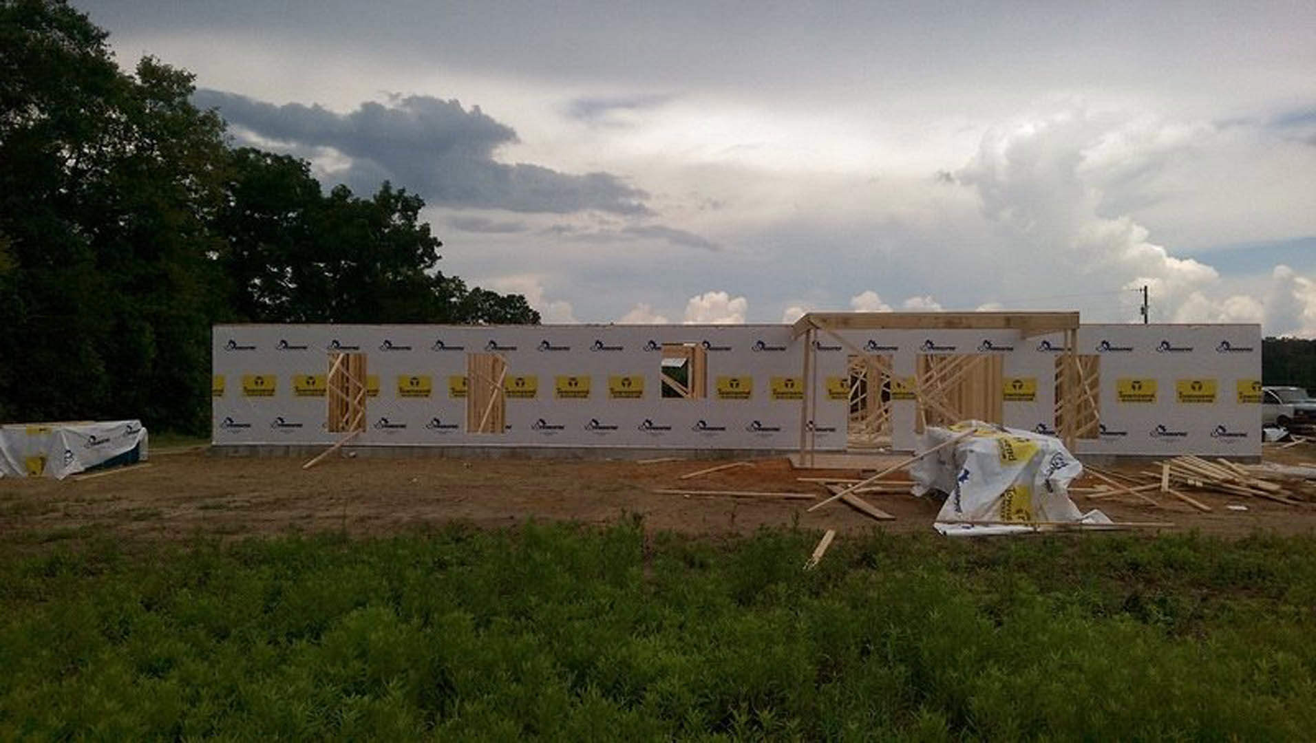 Framed custom home under construction with exposed wood, white board siding, yellow caution signs, patchy green grass and dirt in foreground, cloudy sky and trees in background