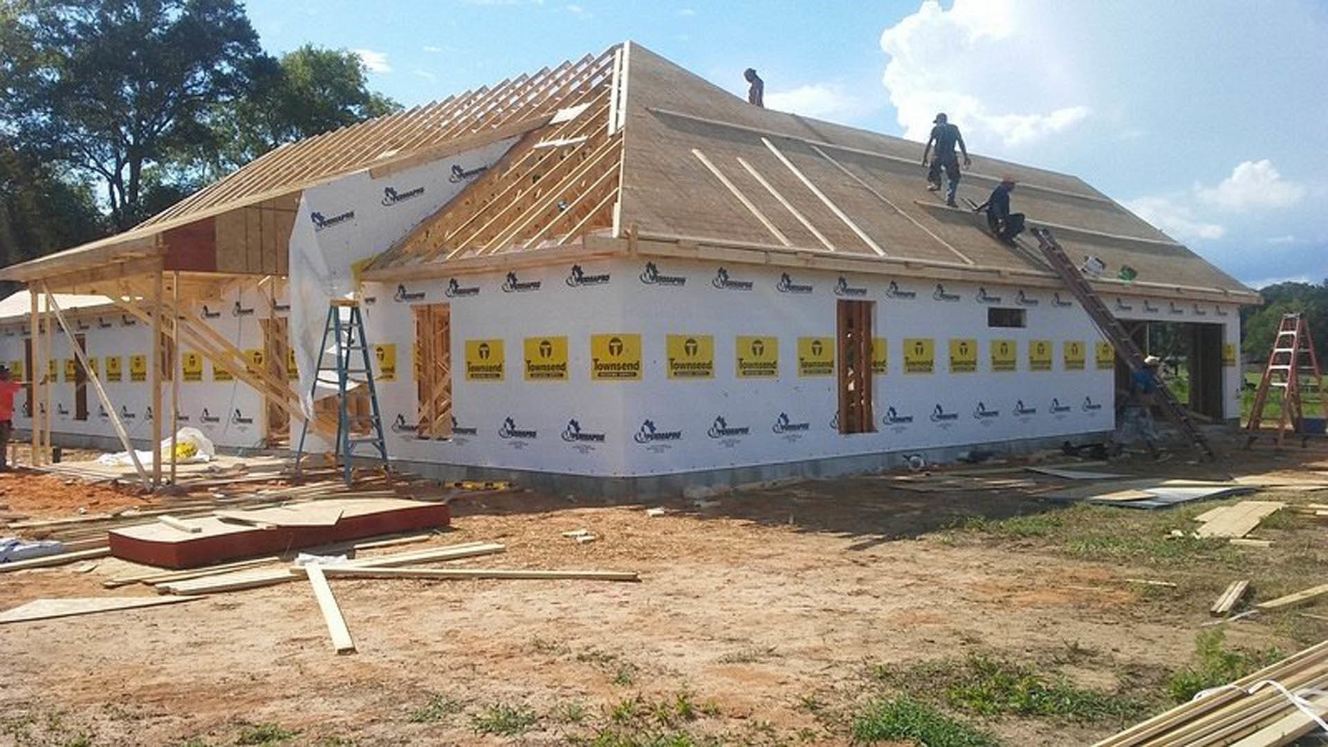 Partially constructed house with exposed wooden framing, workers installing roof panels, scattered lumber in foreground, cloudy sky overhead, trees in background