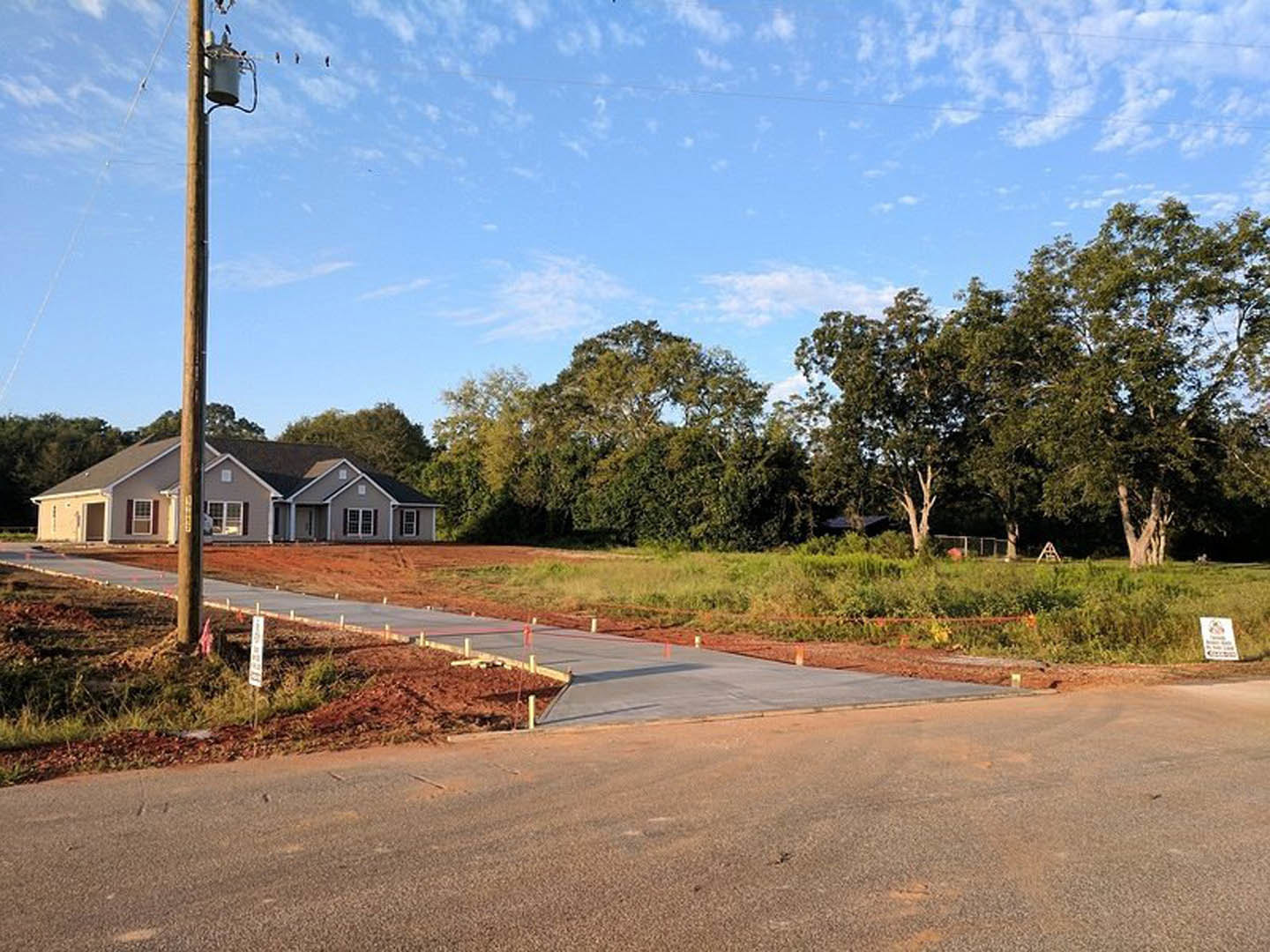 Two-story custom home with light siding, concrete driveway connecting to paved road, mature trees and grass lawn, telephone pole near street, white yard sign on front lawn, partly