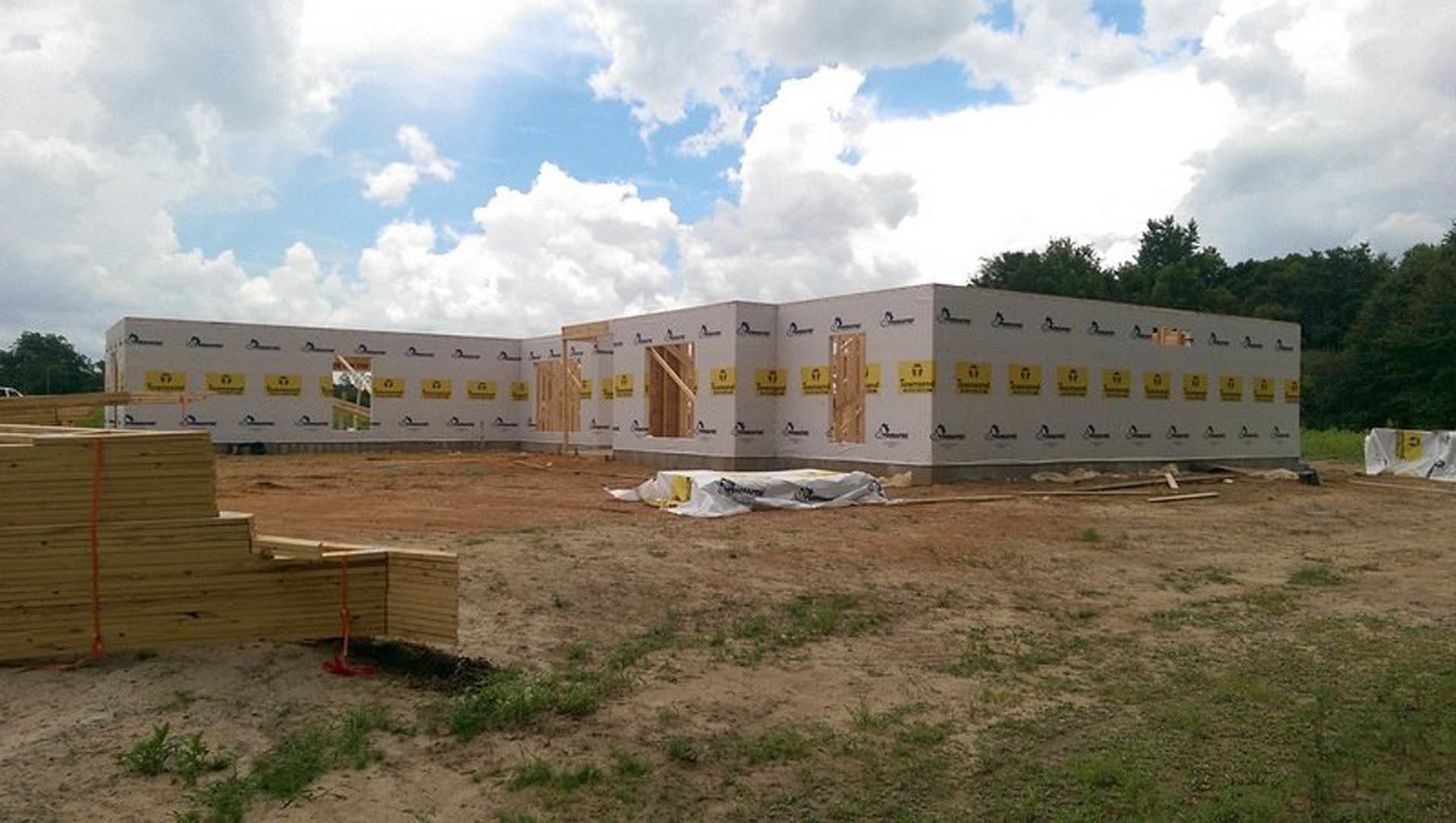 Partially built home with exposed wooden framing, white tarp covering dirt lot, stacks of lumber, and blue sky with scattered clouds