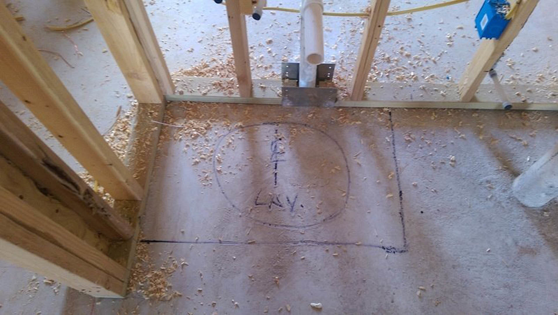 Wood framing with exposed beams and insulation, circular pipe emerging from subfloor, blue electrical box with white label, hand-drawn markings on plywood floor.