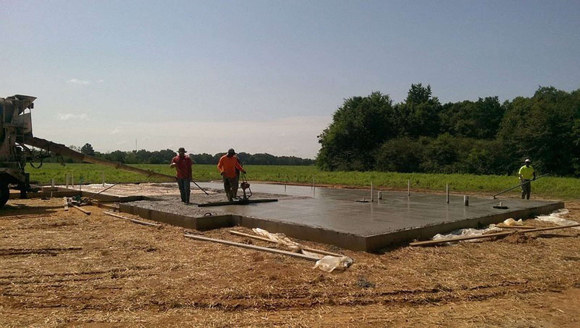 Crew pouring and leveling wet concrete slab foundation outdoors, surrounded by construction equipment, muddy soil, and grassy area with trees in background