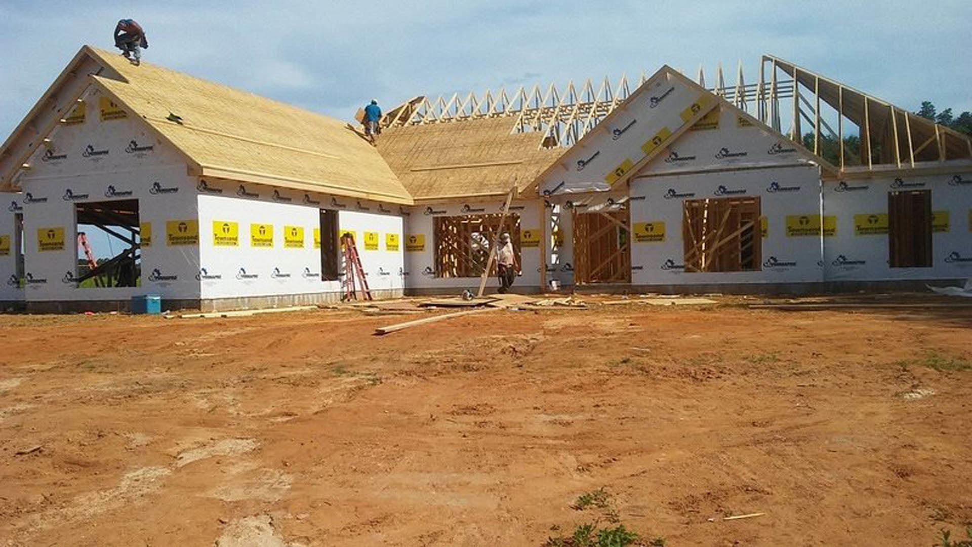 Wood-framed house under construction with two workers on the roof, dirt field in foreground, yellow caution sign, cloudy sky overhead