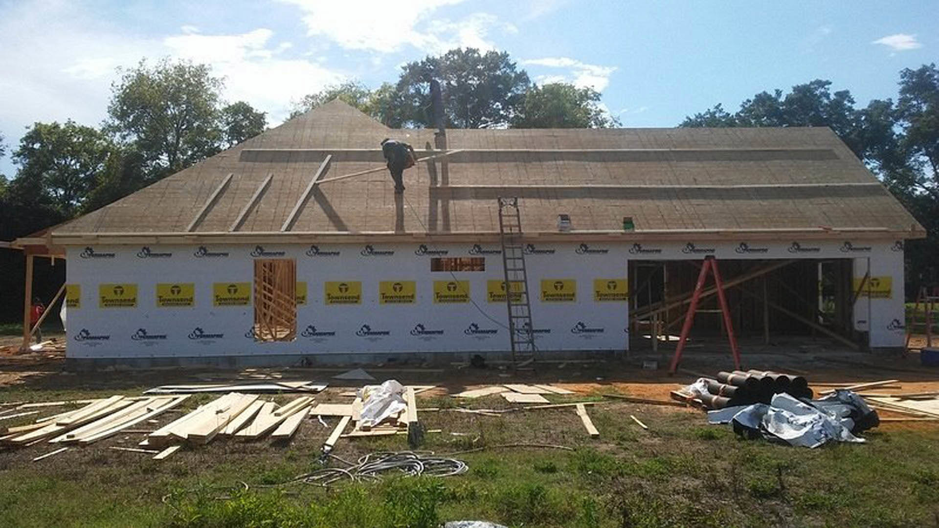 Shingle roof with a man standing near the peak, surrounded by trees and cloudy sky, grass and lumber visible at ground level