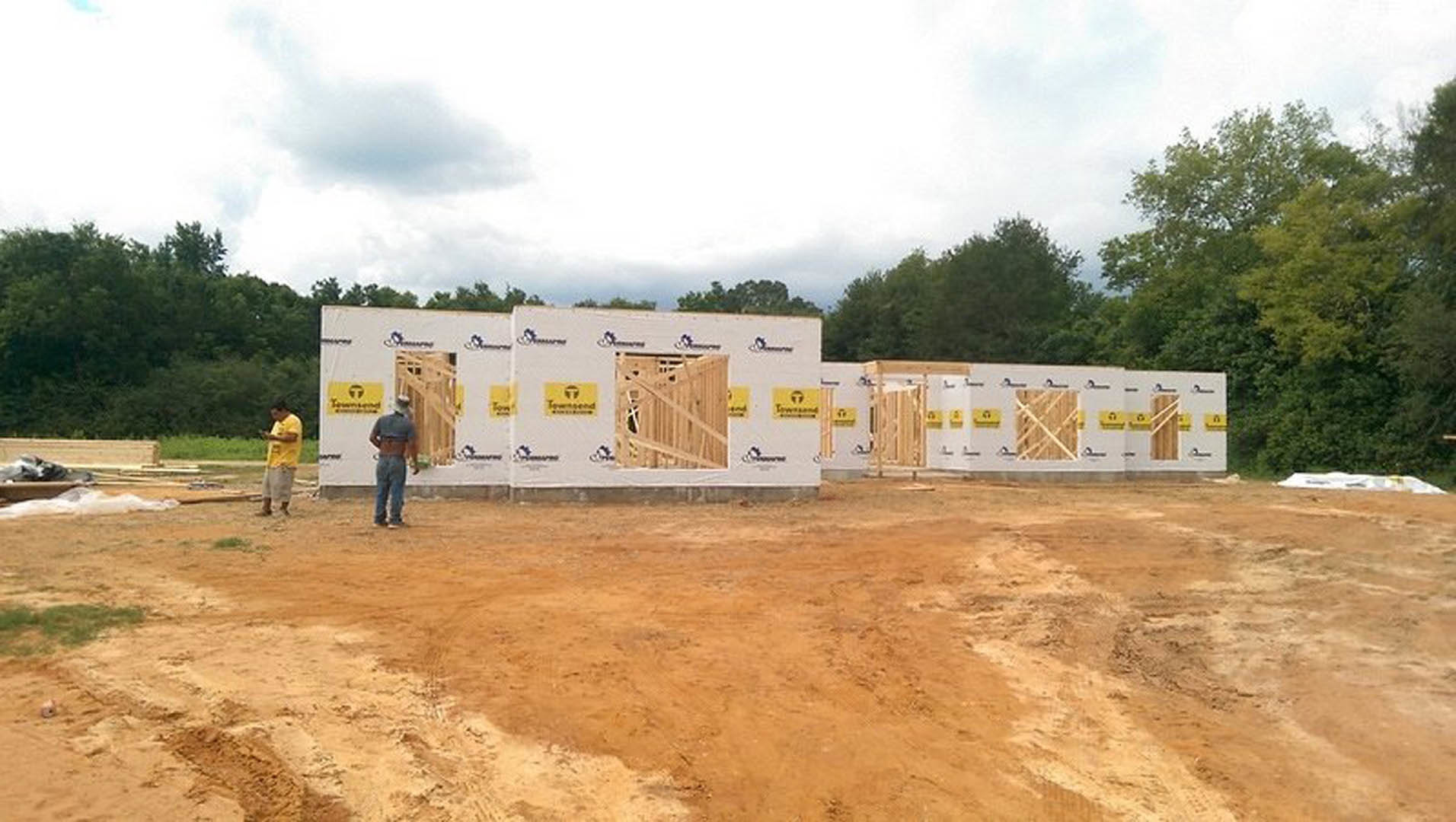 White modern home exterior with yellow construction signs, unfinished landscaping, yellow fire hydrant, and man standing near entrance; cloudy sky and trees in background