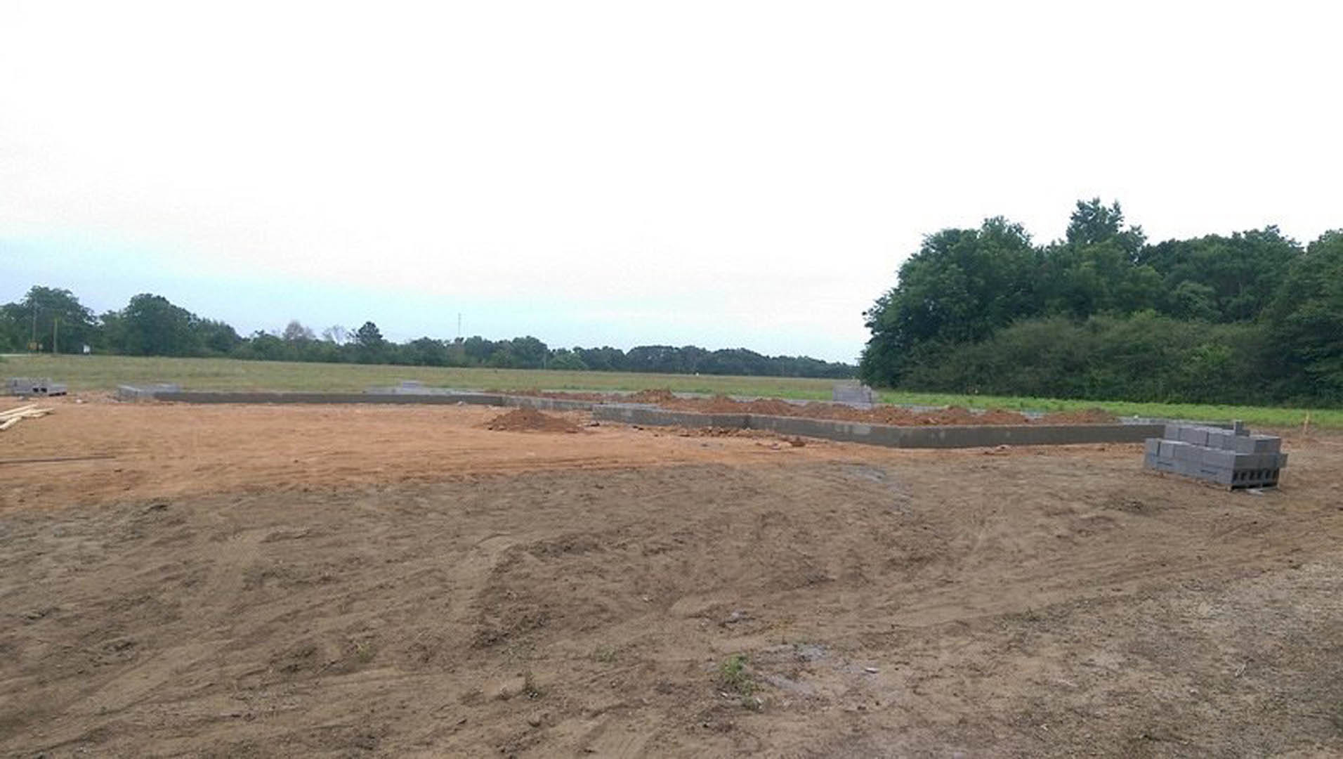 Expansive dirt lot bordered by a concrete wall, scattered trees in the background, muddy ground with patches of grass, overcast sky above