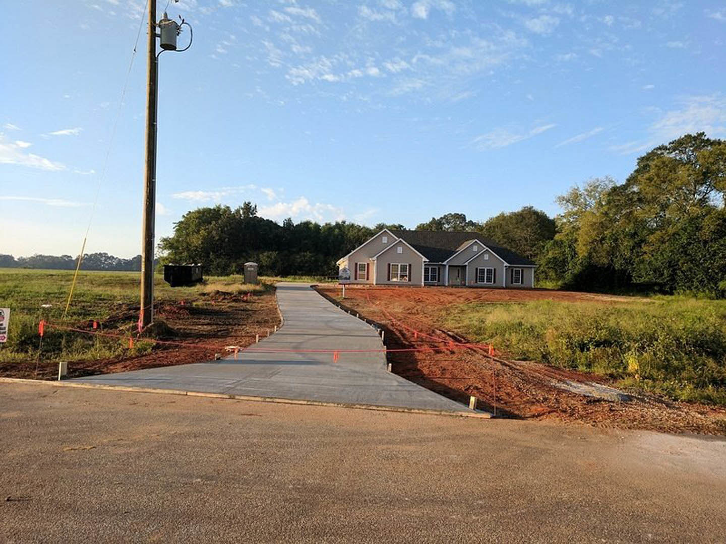Partially built house with black roof, exposed framing, and construction materials, situated beside a concrete road with a telephone pole and red sign; grassy lot and scattered