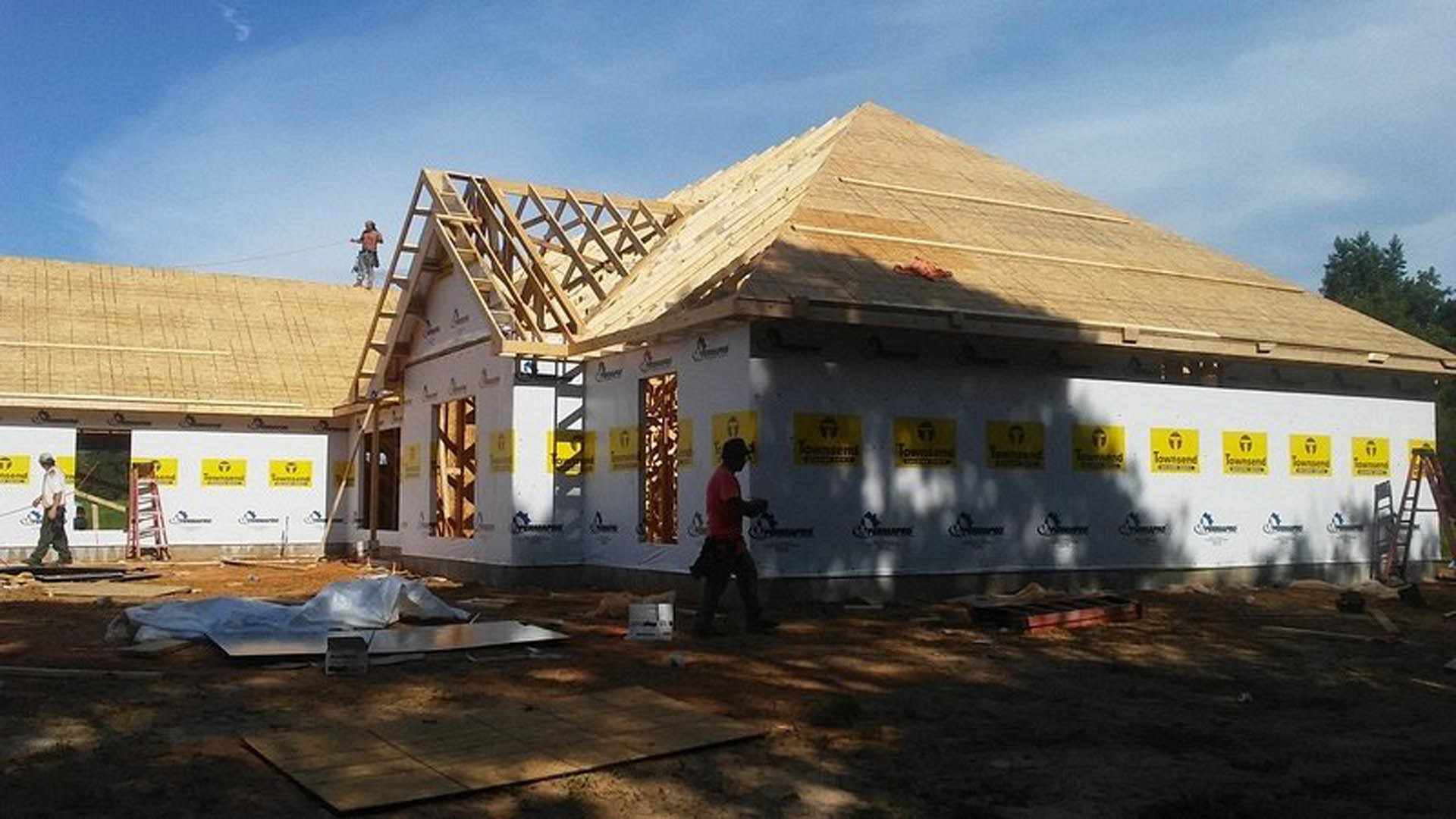 Man in red shirt walking past partially built house with exposed framing, construction materials, and yellow caution sign on metal pole; cloudy sky overhead.