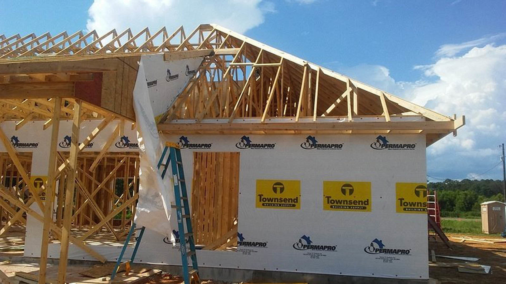 Wooden house frame under construction with a ladder leaning against an exterior wall, surrounded by lumber and open sky.