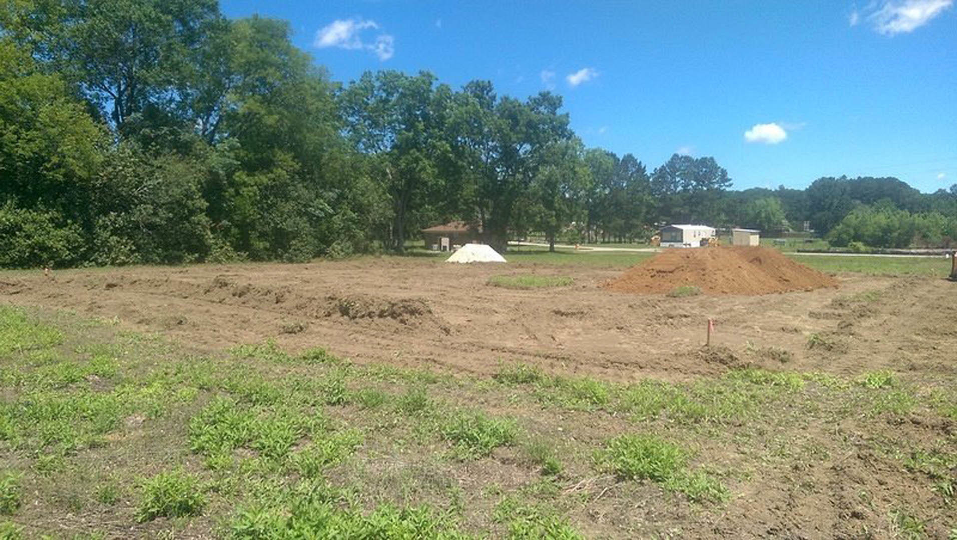 Dirt field with scattered piles of soil, trees lining the background, white building partially visible behind a fence, cloudy sky overhead