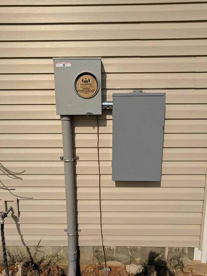 Grey electrical meter box mounted on exterior wall with white pipe and black cable visible.