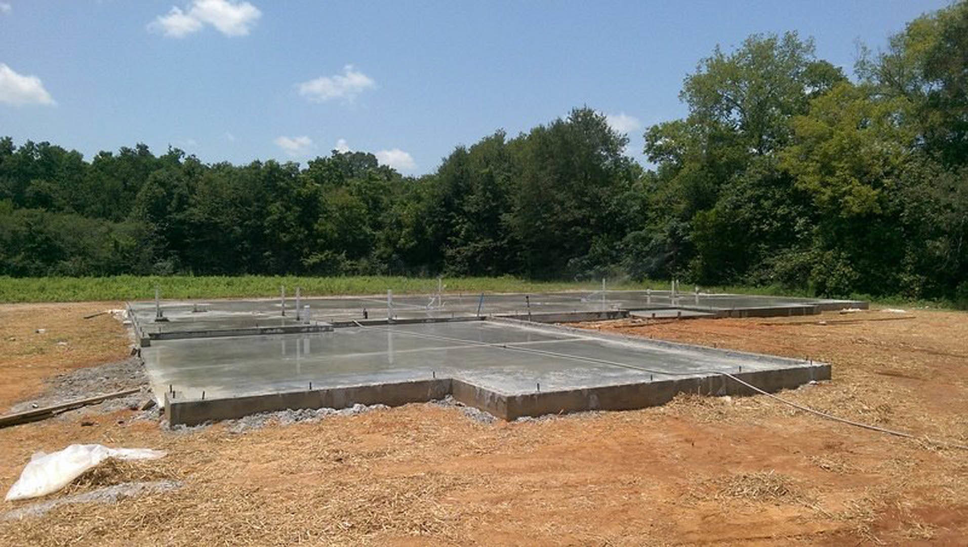 Concrete slab foundation with square layout in grassy field, white bag on ground, surrounding trees, blue sky with scattered clouds