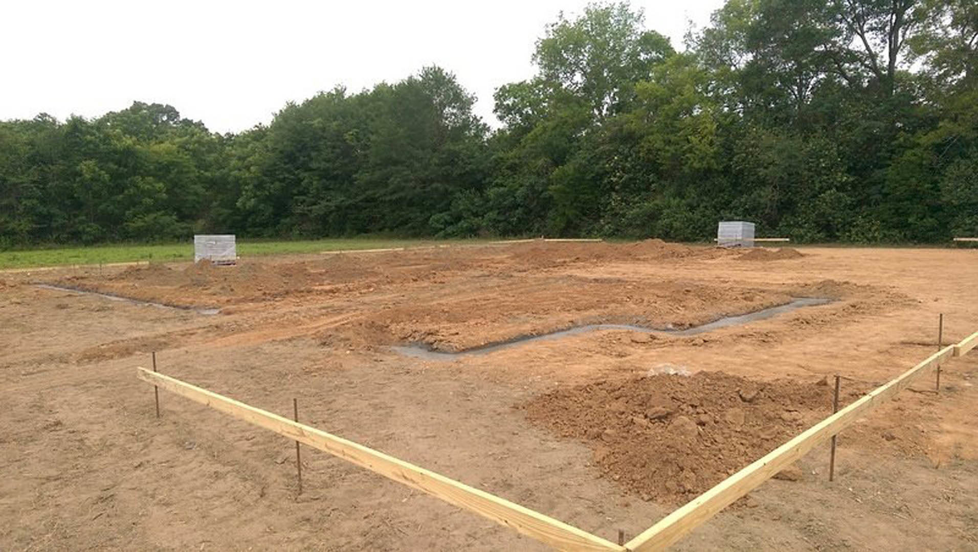 Dirt lot bordered by a wooden fence, mature trees in the background, wooden boards and framing scattered across soil