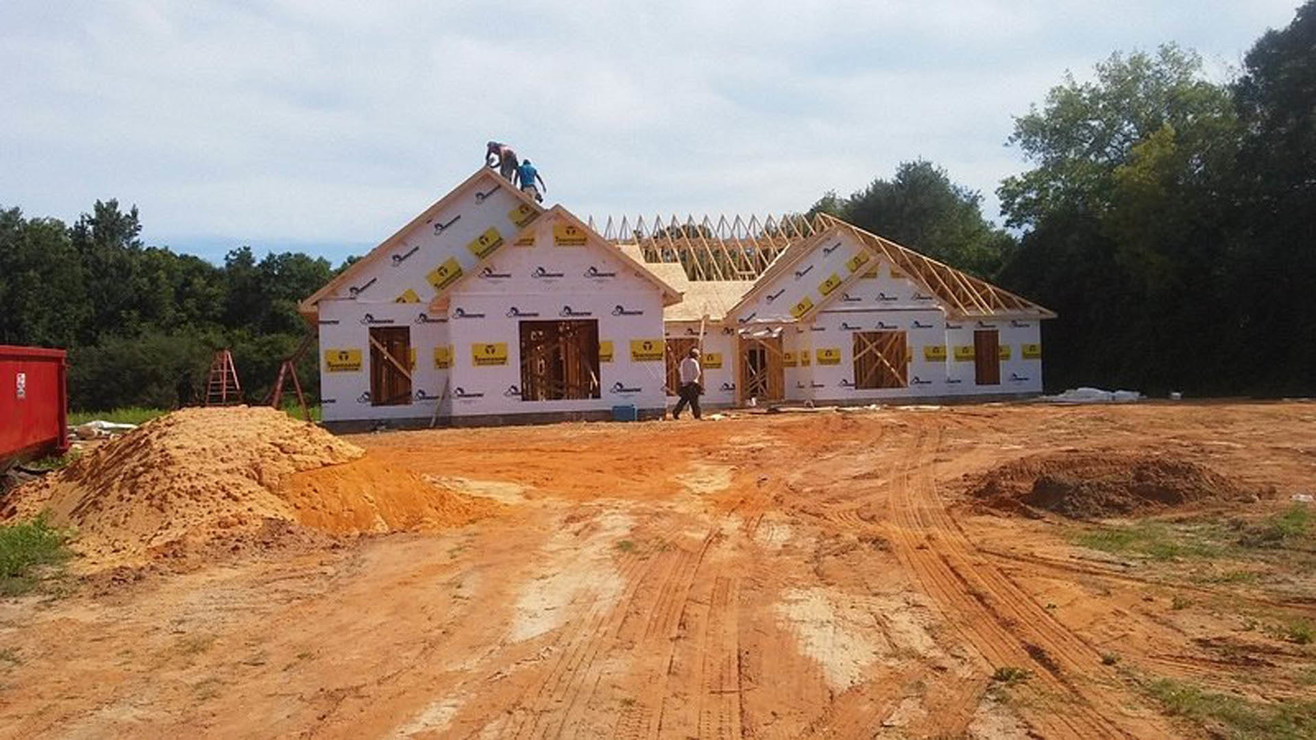 Partially built house with exposed concrete walls, workers installing roofing materials, piles of sand and construction debris in foreground, trees and cloudy sky in background