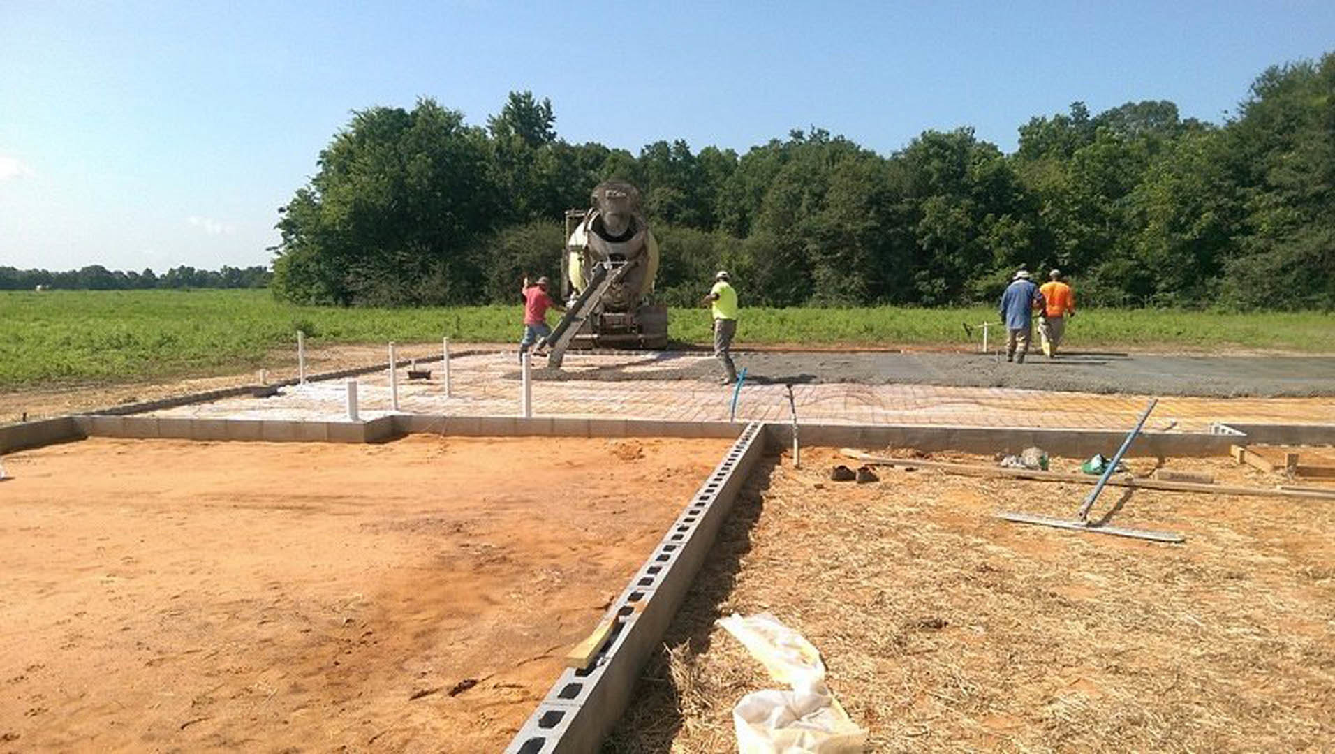 Group of people gathered near a partially built concrete foundation outdoors, surrounded by soil, grass, and trees under a cloudy sky