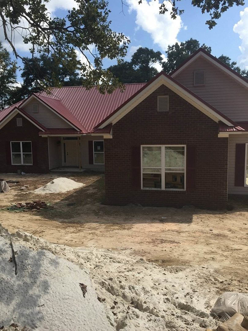Partially built house with red roof, white framed windows, dark brown door, white door with black handle, surrounded by dirt, white rocks, and trees under cloudy sky