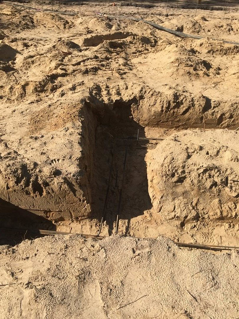 Deep hole dug into sandy ground, surrounded by loose dirt and small rocks, with a fence visible in the background.