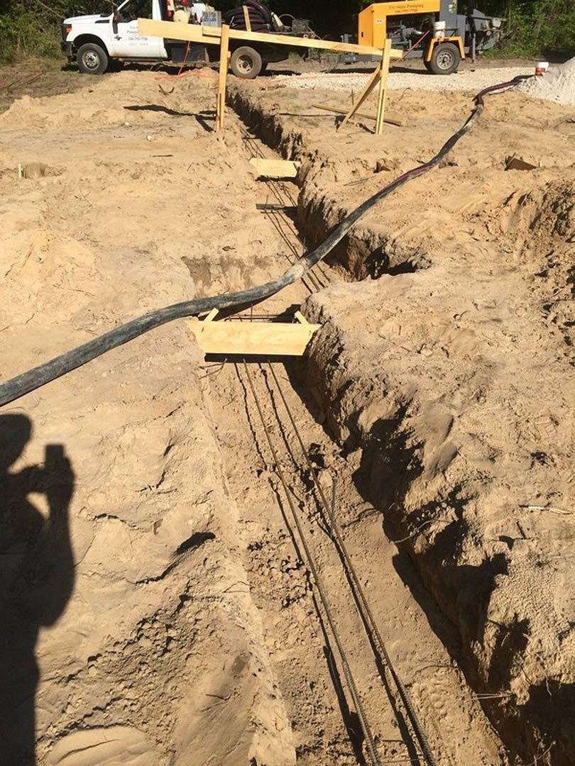 Excavated trench in sandy soil with exposed pipe and electrical wires, shadow of a person’s face cast across ground, nearby trailer with wooden frame visible in background