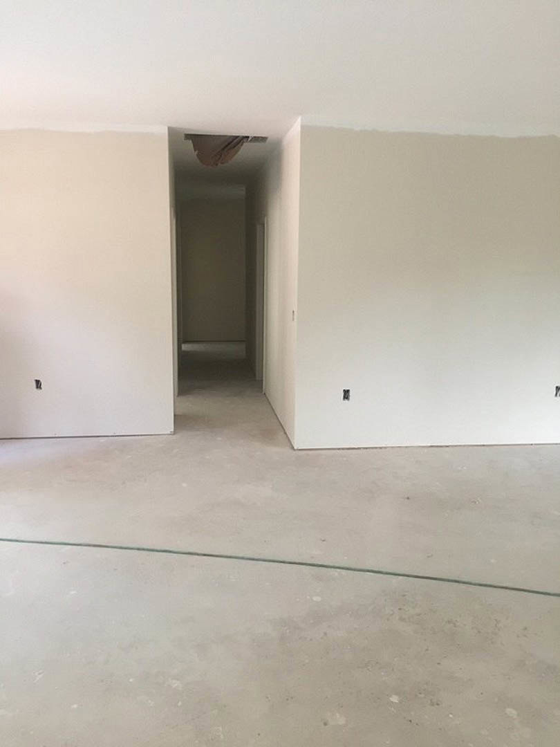 White plaster wall with a rectangular hole, concrete floor showing a visible crack, hallway featuring a ceiling fan, and a white door with matching frame; black border accent along