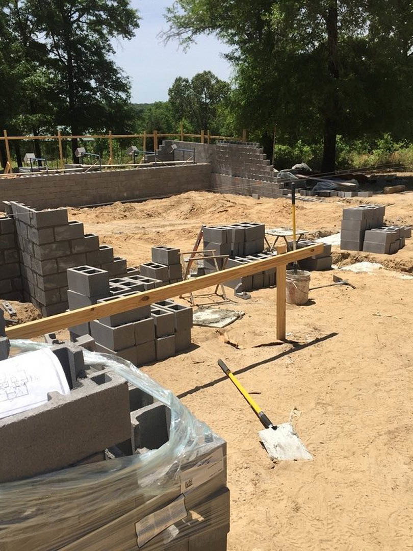 Construction site with stacked grey bricks, metal shovel resting on soil, bucket with handle, close-up blueprint, yellow and black pencil, exposed foundation, outdoor setting with