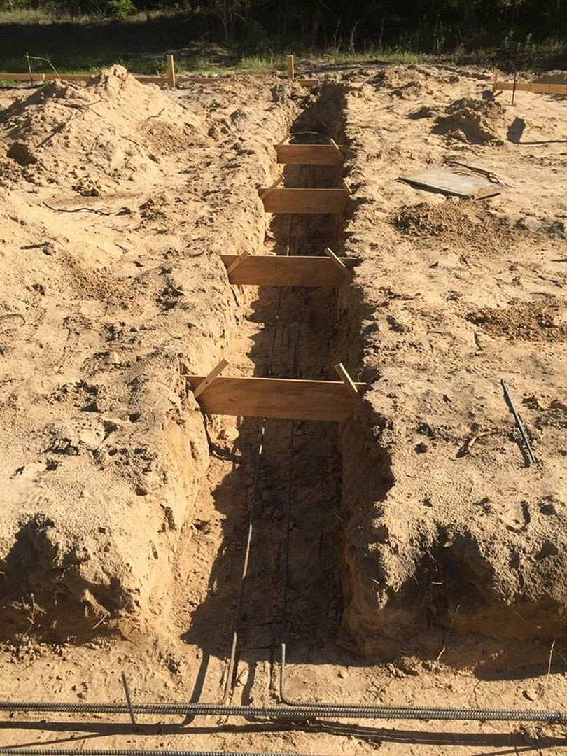 Narrow trench dug in sandy soil with exposed rocks, wooden board illuminated by sunlight, wire visible in foreground, wooden post standing in open field