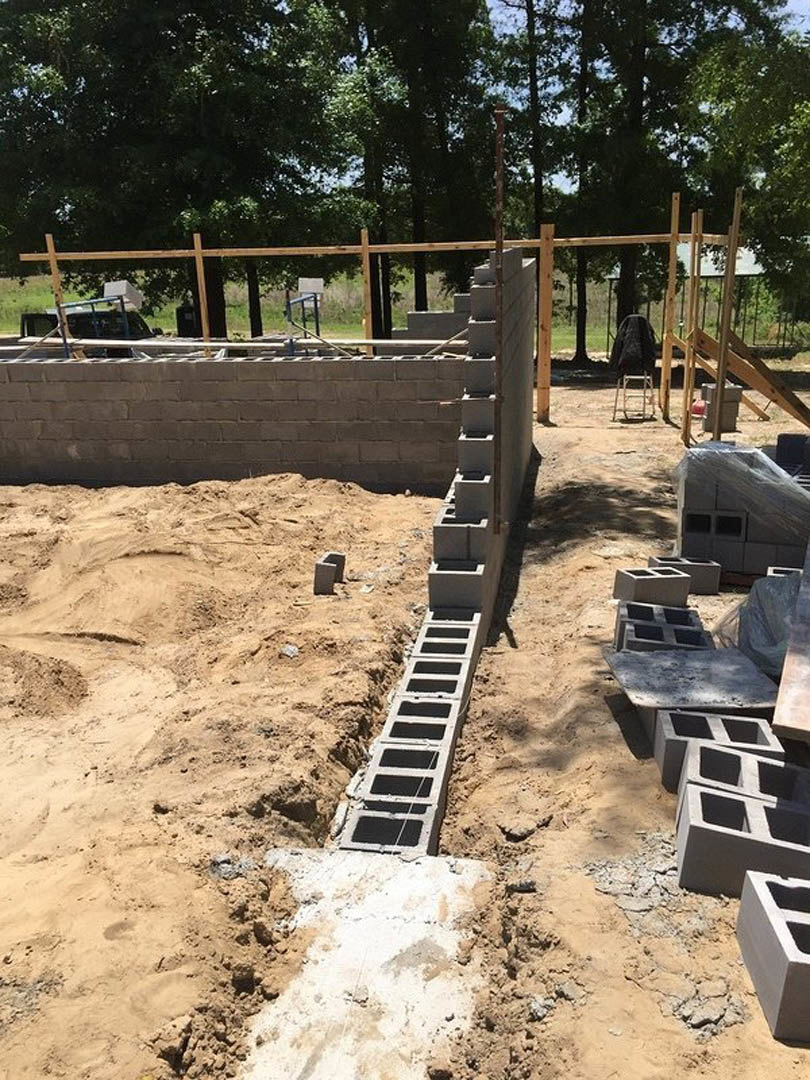 Stacked concrete blocks wrapped in plastic beside a metal fence on a soil foundation, surrounded by trees and plants at an outdoor construction site.