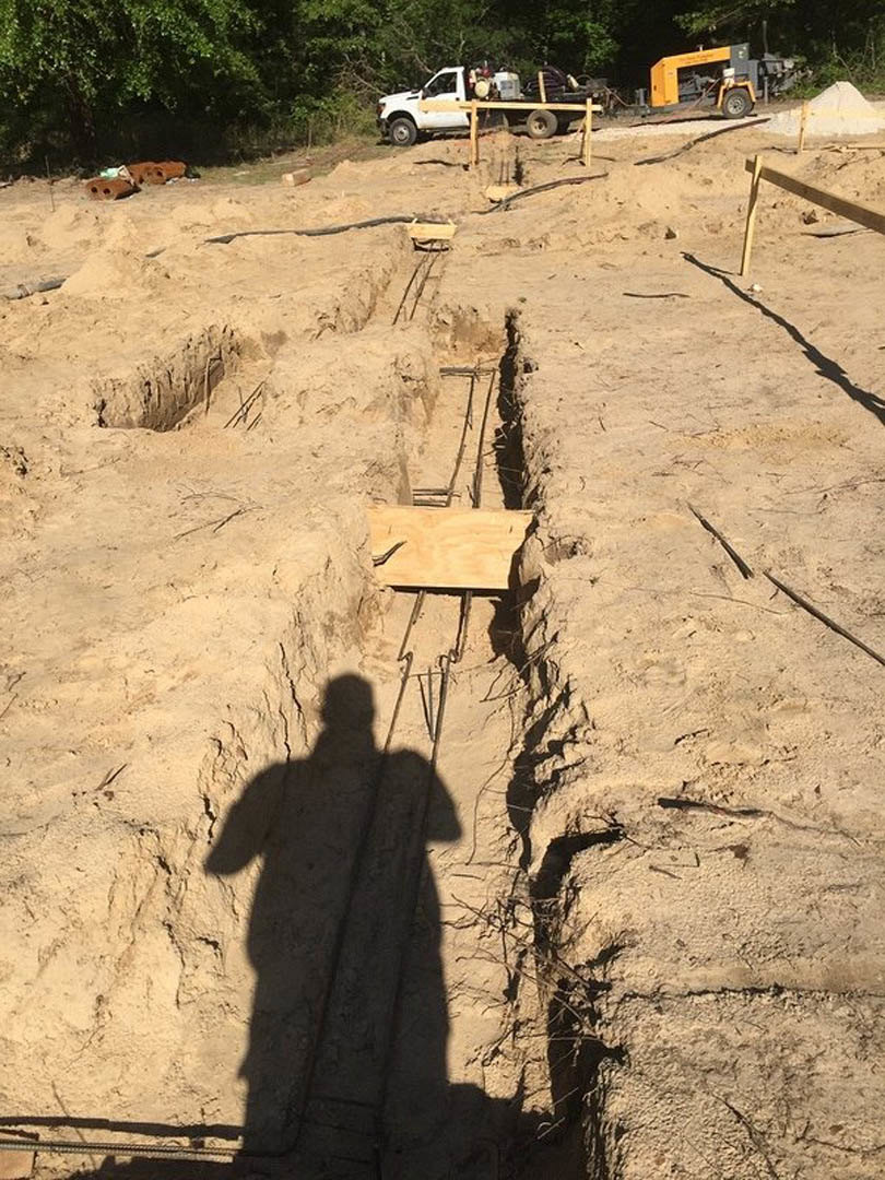 Shadow of a person cast across a dirt field with a white truck and tractor in the background, sandy soil and a white pyramid-shaped object nearby