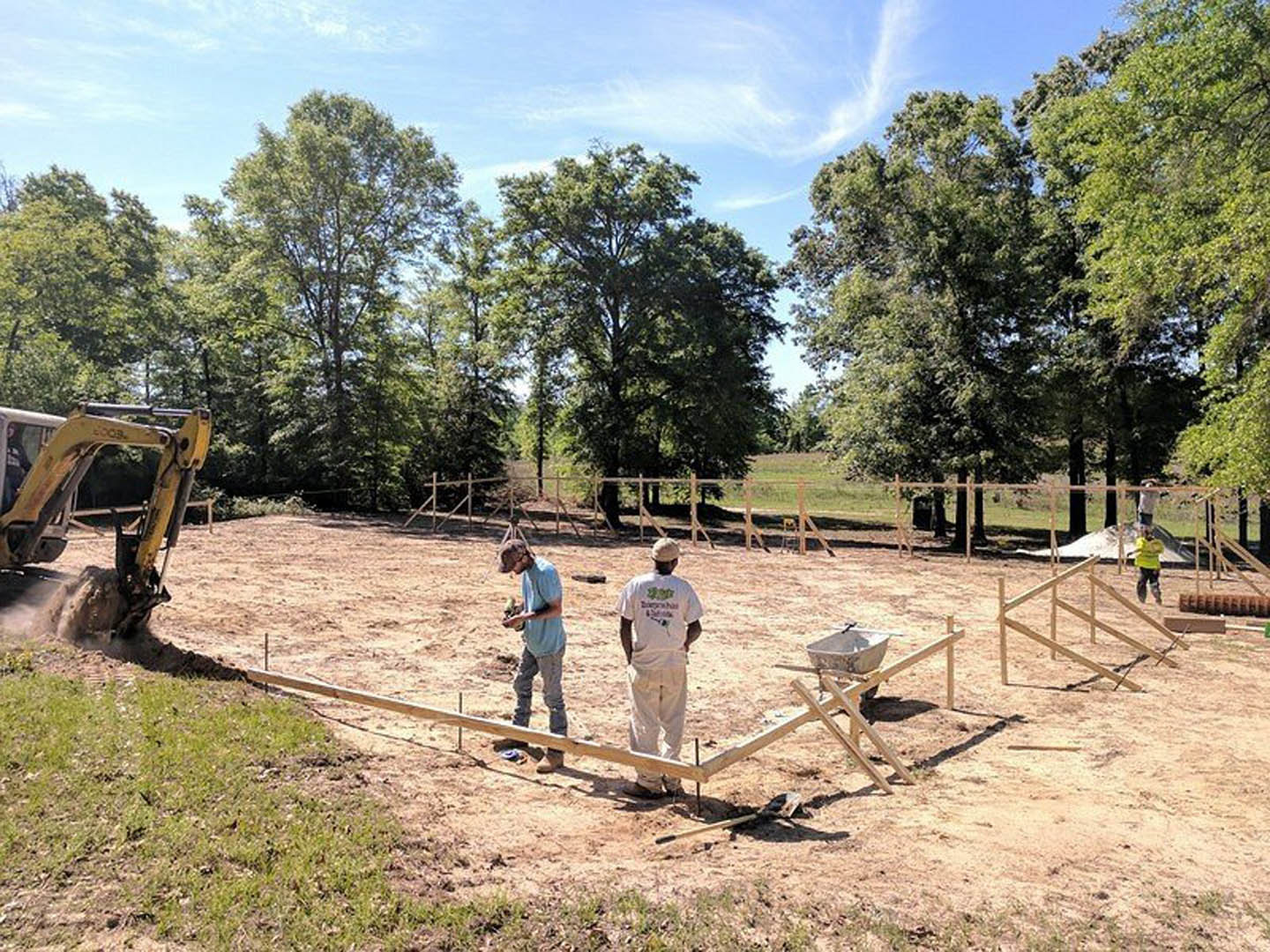 Two men standing on bare soil near construction equipment, surrounded by trees and open sky