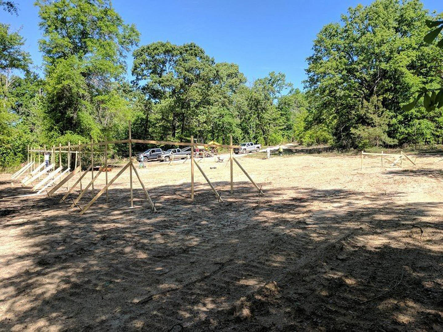 Dirt field bordered by a wooden fence, clusters of leafy trees under clear blue sky, sparse vegetation on ground