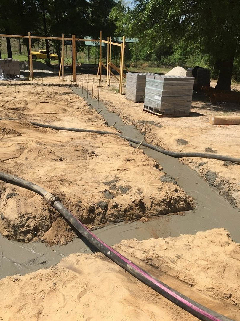 Concrete foundation with exposed pipes, stacked bricks on pallets, plastic-wrapped materials, yellow truck parked near fenced area, surrounding soil and scattered construction