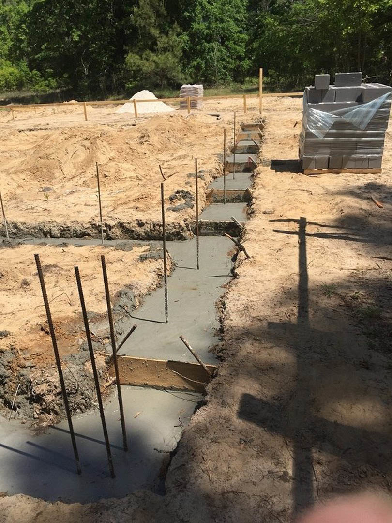Concrete piles and stacks of grey bricks wrapped in plastic on a muddy construction site, with a metal fence in the foreground and trees in the background.