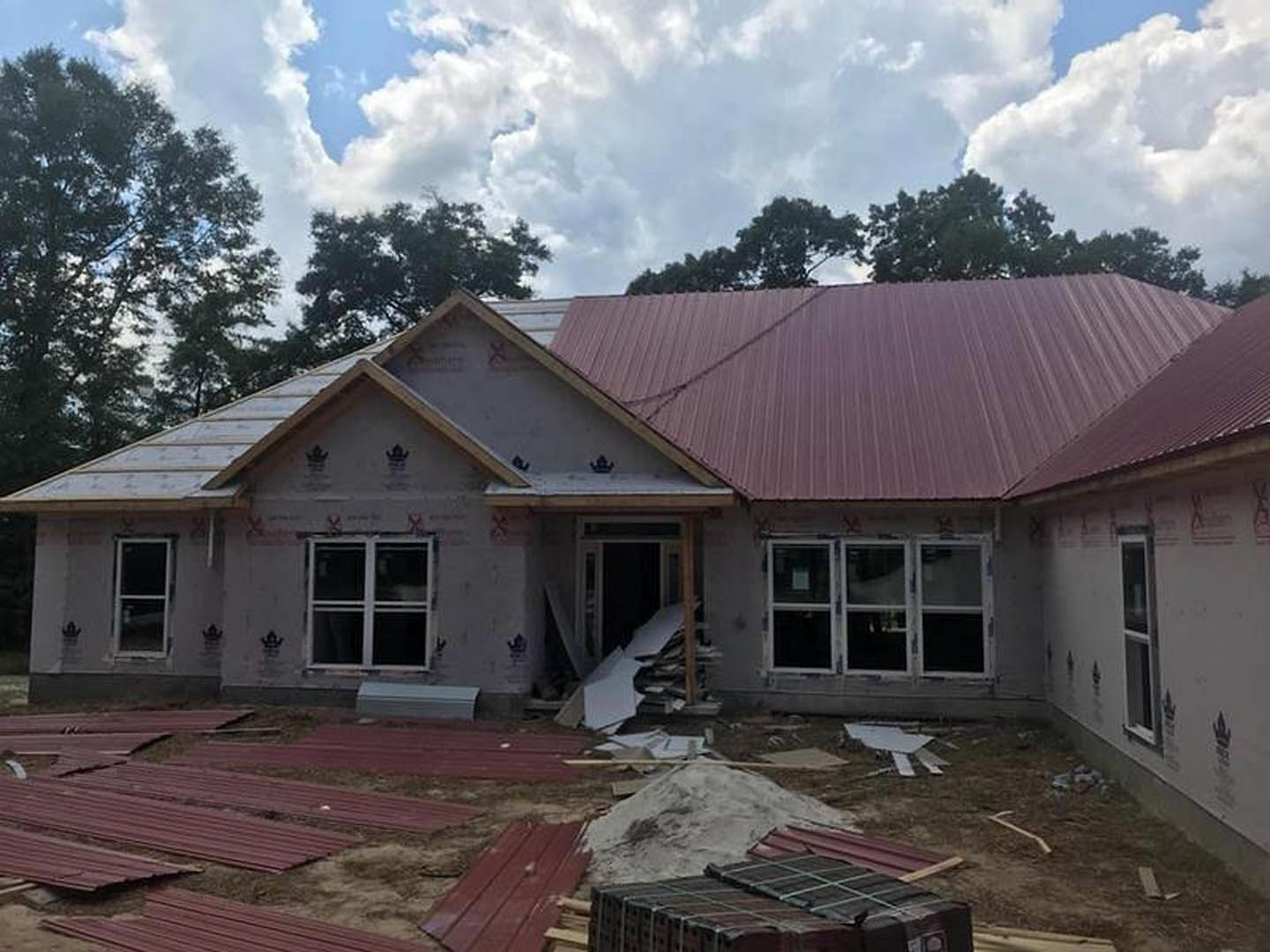 Red metal roof atop partially built house, white-framed window, construction debris scattered in front, cloudy sky overhead.